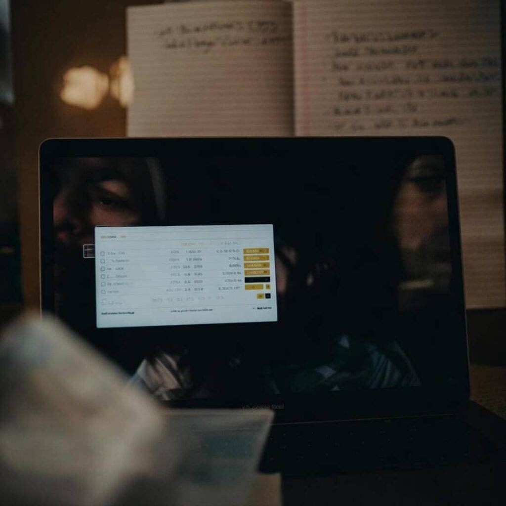 Laptop screen reflects tired face, messy notes.